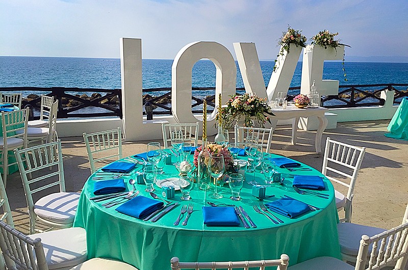 An outdoor wedding reception with round tables decorated with turquoise table clothes and blue napkins, with the word "Love" in large and the ocean behind it.