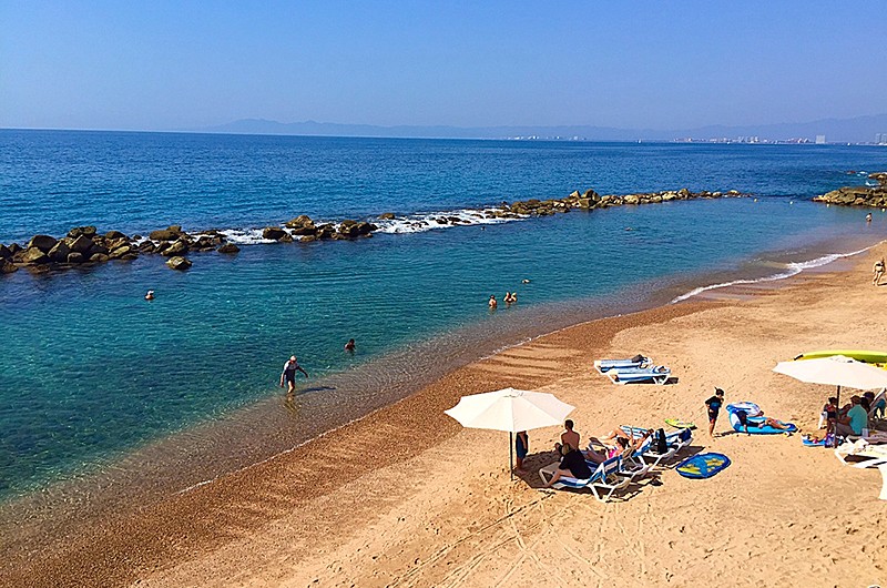 People enjoying the beach with lounge chairs and two umbrellas while people play in the water.