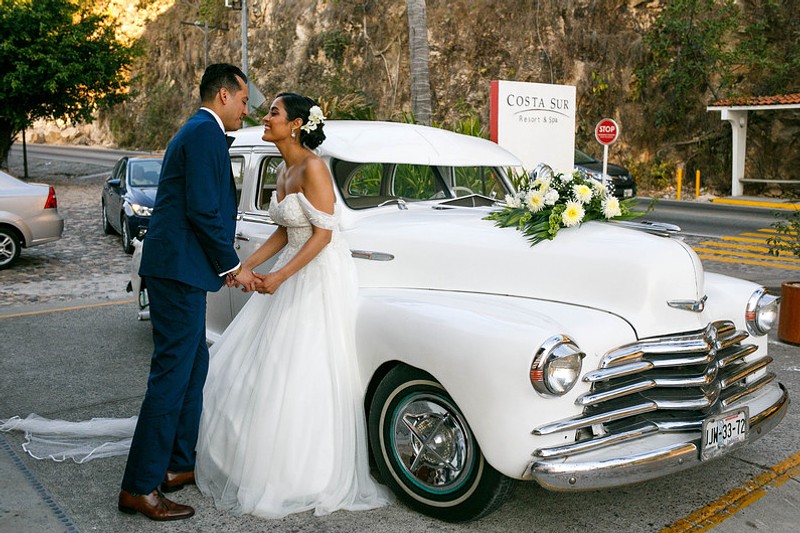 Bridal photo of couple leaning against a vintage A white 1940s-era car adorned with a floral arrangement parked in front of the resort.