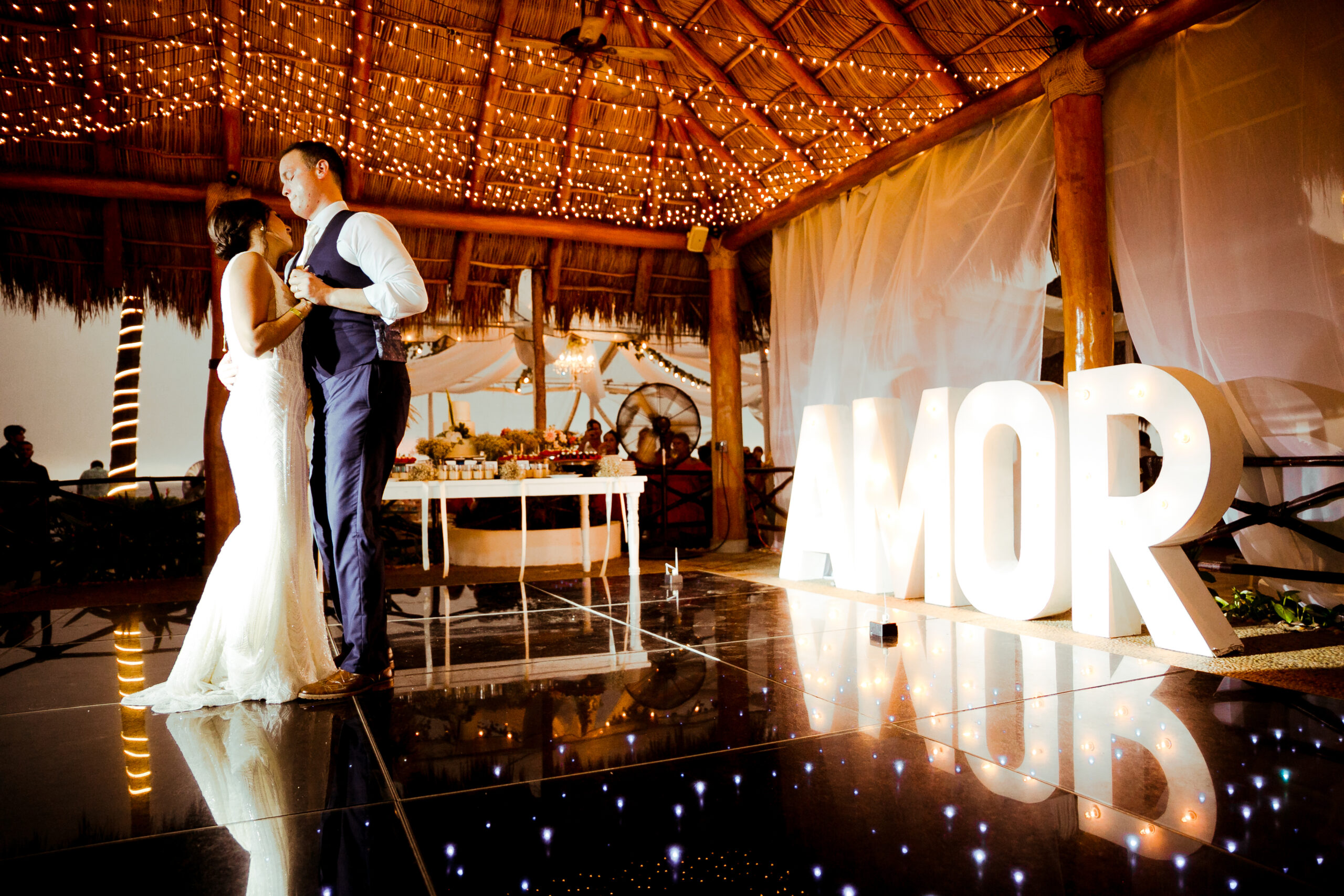 A couple slow dancing on a reflective dance floor at a wedding reception. The word "AMOR" is displayed in large white letters behind them.