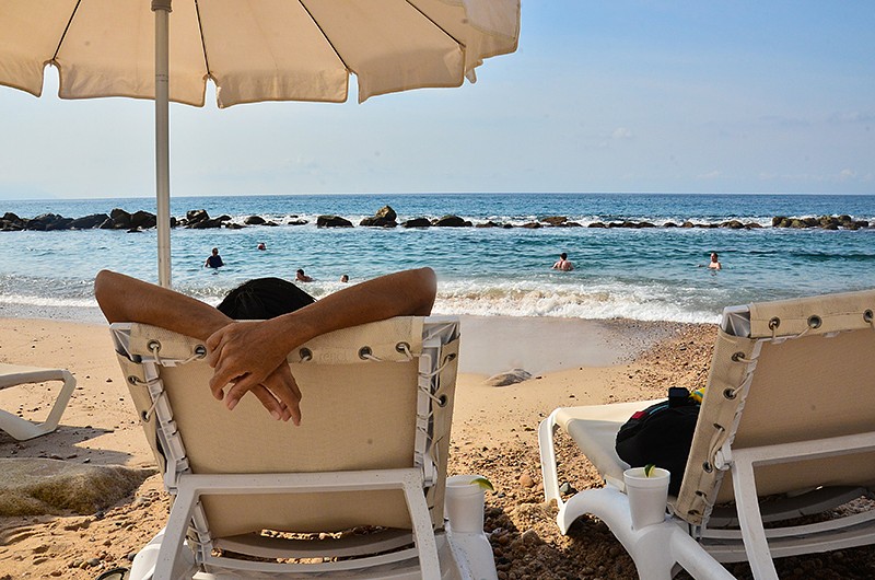 Perspective view of a person relaxing on a lounge chair looking out to people playing in the ocean.