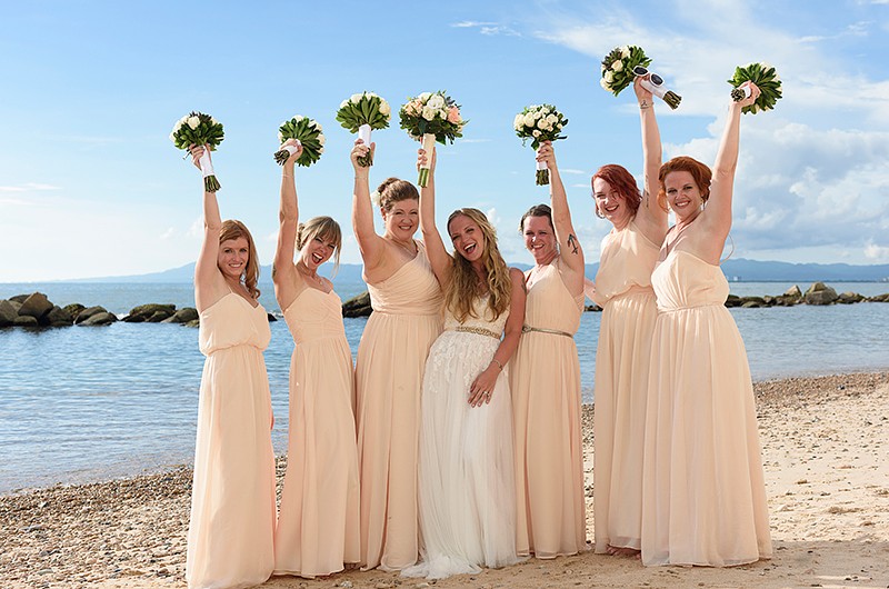 Bride poses with her bridesmaids wearing beige. They are smiling and are all raising their bouquets in the air.