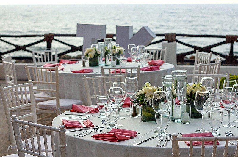 A wedding reception with round tables with red napkins and white wooden chairs. The letters "T" and "K" in large objects behind the tables overlooking the ocean.