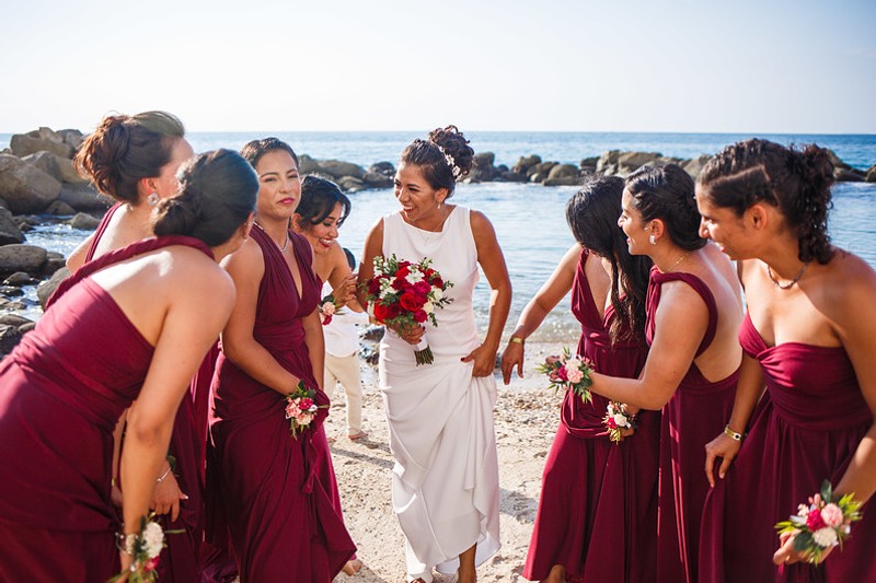 A bride in a white dress holding a bouquet of red and white flowers gathered with her brides maids wearing red dresses on the beach.