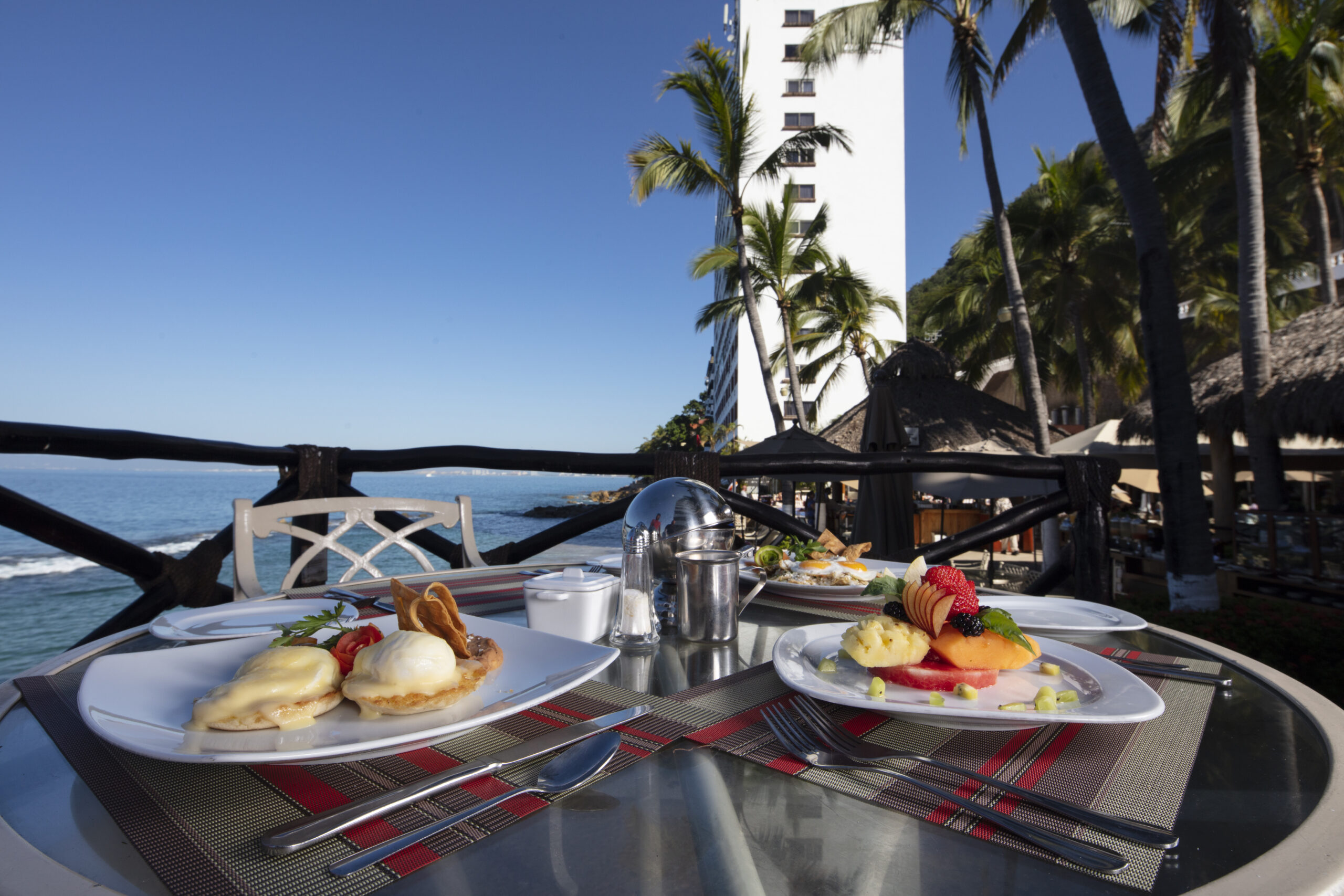 A table on a balcony overlooking the ocean has two plates of breakfast food. One plate has eggs Benedict, and the other has fruit and pancakes. There are palm trees and a hotel in the background.