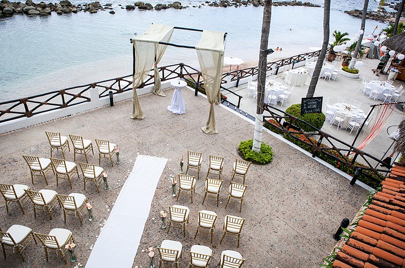 Aerial view of wedding ceremony, styled with white aisle carpet and alter decorations. A row of wooden chairs with white cushions line the aisle.