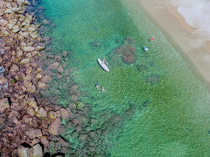 Aerial view of a person paddle boarding a four people swimming in clear water near the rock jetty.