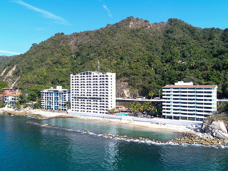 View of three hotel buildings in front of the beach with mountains behind them.