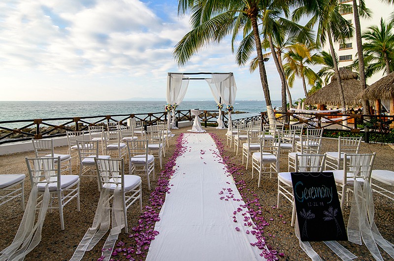 A beach wedding ceremony setup with a pink with a cabana style white archeway, white chairs decorated with white sashes. The ocean and palm trees are visible in the background.