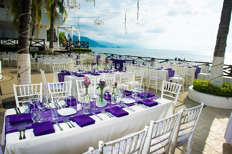 An outdoor wedding reception with rectangle tables, string lights, and stunning ocean views. The decorations are purple colored and tables and chairs are white.