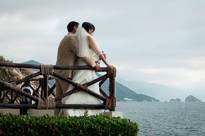 A photo of a bride and groom standing on a balcony overlooking a beautiful ocean view. The bride is wearing a white dress and the groom is wearing a tan suit. They are holding hands and looking out at the view.