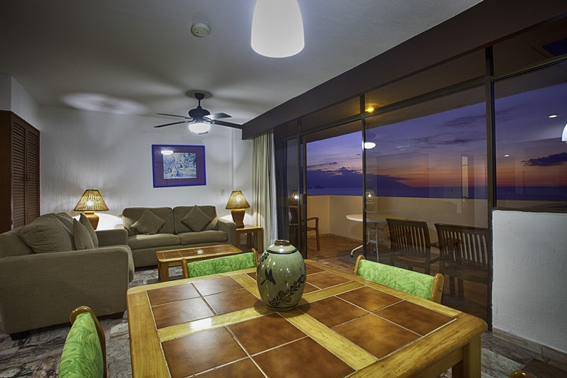 View of a room's seating area with two couches, coffee table and dining table overlooking the balcony with sunset views.