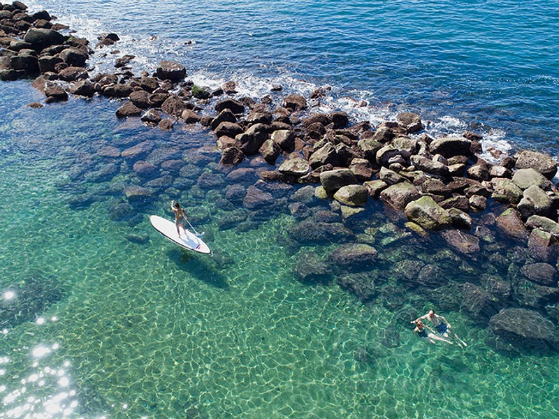 Person paddle boarding near two swimmers in clear water.