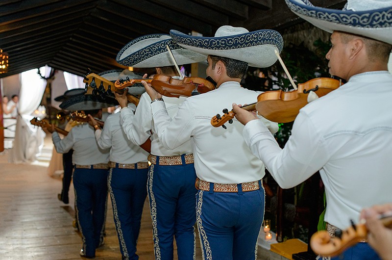 A mariachi band, dressed in white dress shirts, blue pants with designs, and white sombreros with blue decorations, walks into a venue playing violins.