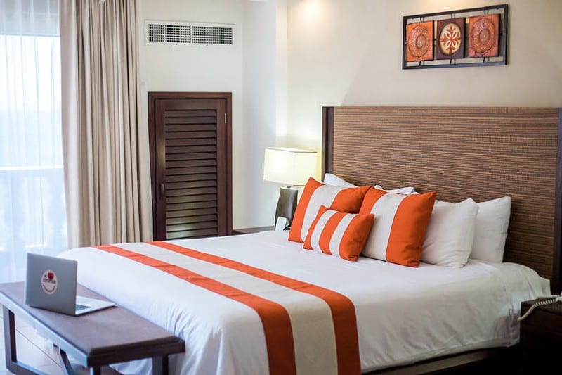 Single bed with orange colored decorative pillows and cover, bench in front of bed with a laptop on top.