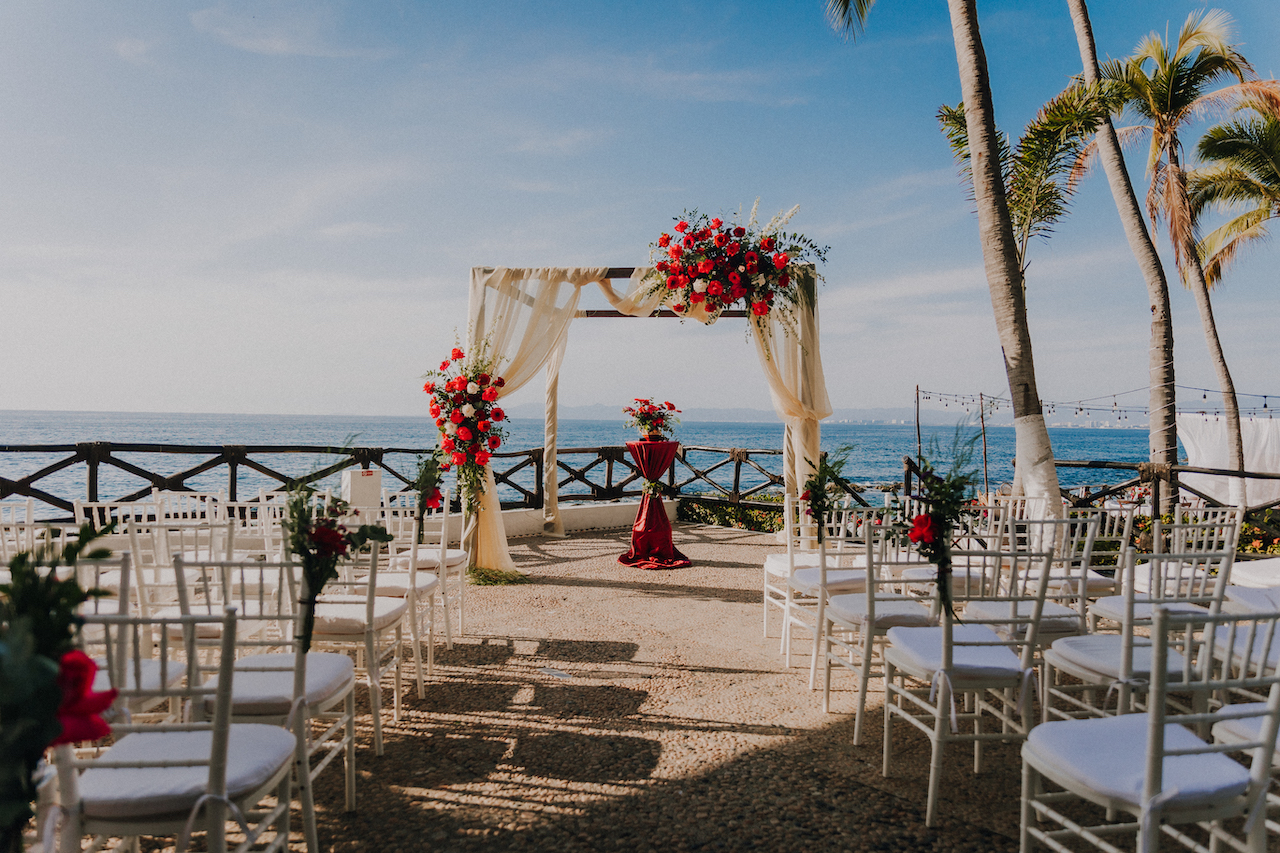 A beach wedding ceremony setup with a red and white floral archway, white chairs, and a red runner on the altar. The ocean and palm trees are visible in the background.