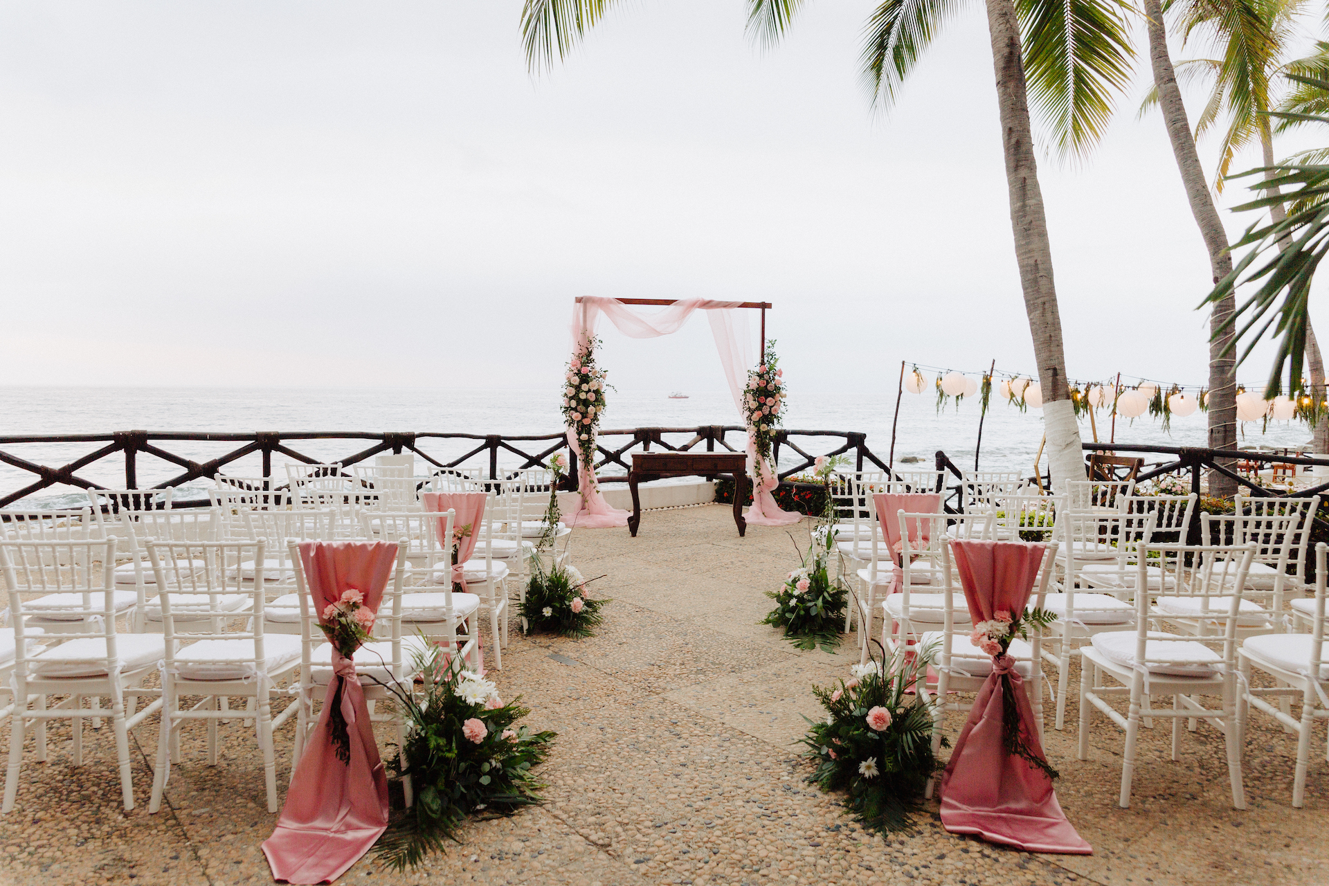 A beach wedding ceremony setup with a pink floral archway, white chairs decorated with pink sashes, and small floral arrangements. The ocean and palm trees are visible in the background.