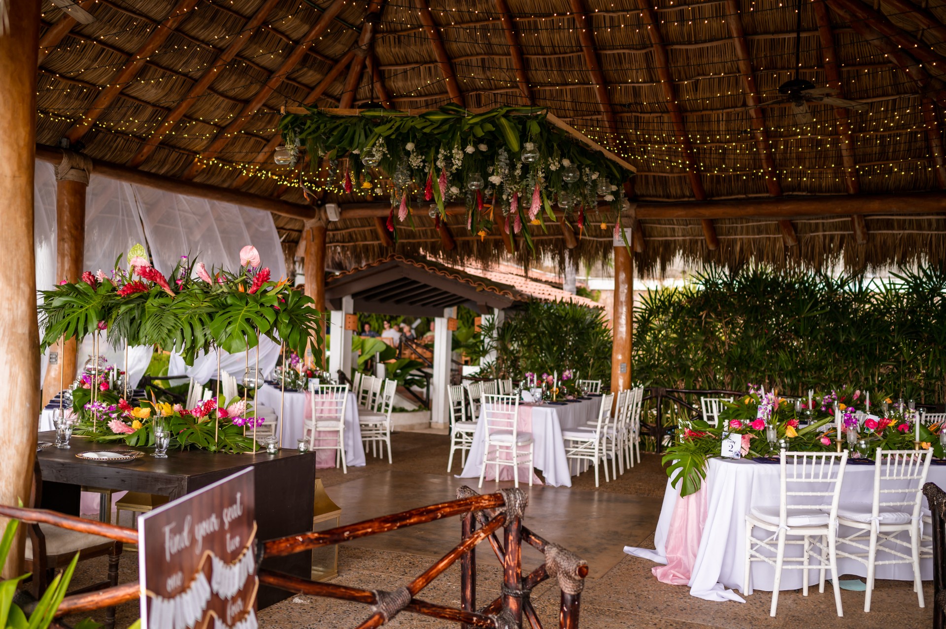 A decorated outdoor wedding venue with hanging floral arrangements, string lights, and tropical foliage.